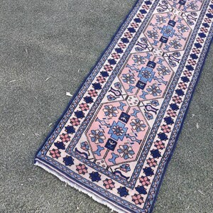 May include: A pink and blue patterned runner rug with a geometric design. The rug has a fringed edge and is made of wool.
