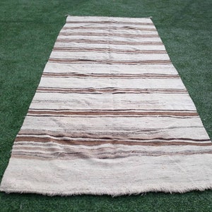 May include: A white and brown striped rug with a fringed edge. The rug is laid out on a green artificial grass surface.
