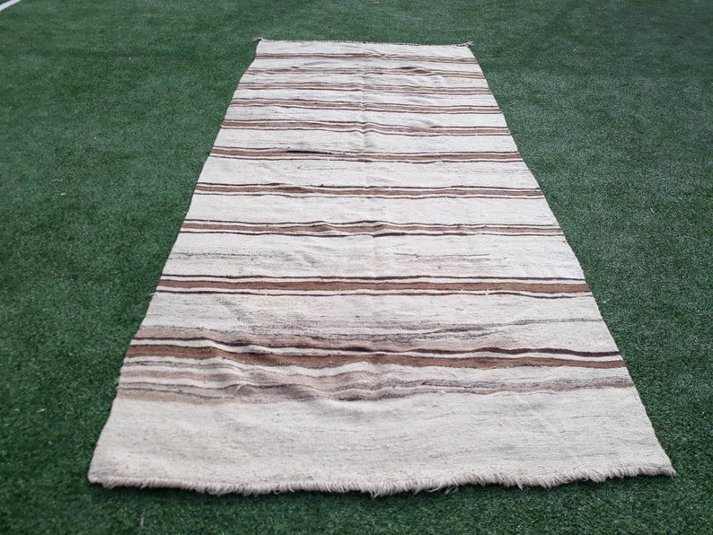 May include: A white and brown striped rug with a fringed edge. The rug is laid out on a green artificial grass surface.