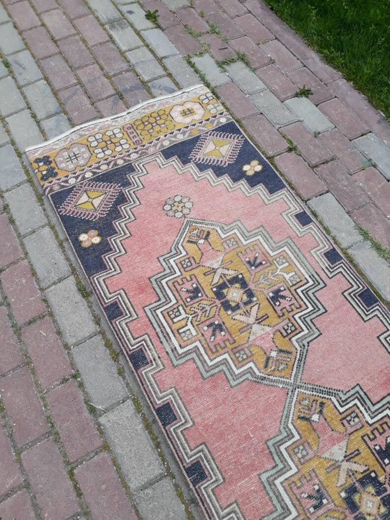 May include: A pink and yellow patterned runner rug with a geometric design. The rug has a black border and is made of a woven fabric.