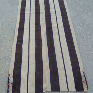 May include: A long, cream-colored rug with dark brown and purple stripes. The rug is woven with a simple pattern and has a slightly frayed edge.