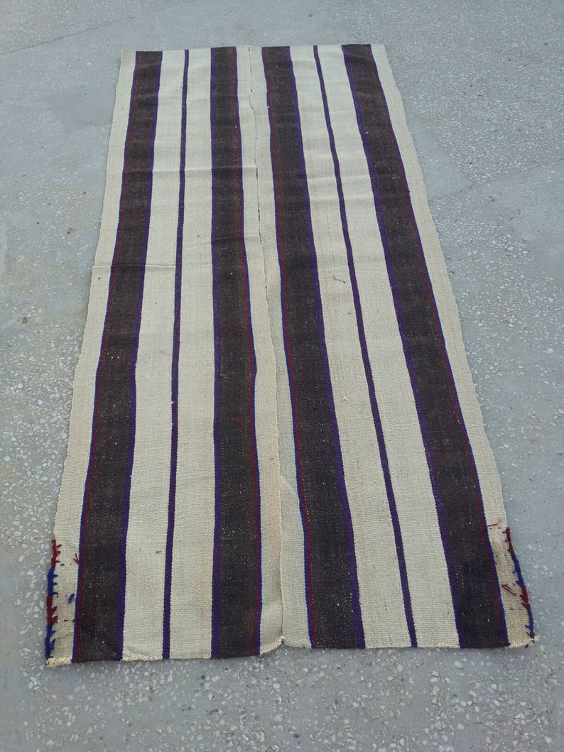 May include: A long, cream-colored rug with dark brown and purple stripes. The rug is woven with a simple pattern and has a slightly frayed edge.