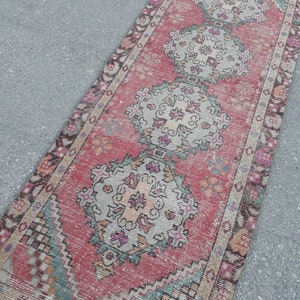 May include: A vintage Turkish runner rug with a faded pink background and intricate floral patterns in shades of beige, green, and brown. The rug has a distressed look and is perfect for adding a touch of bohemian charm to any room.