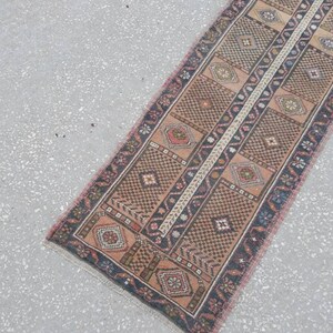 May include: A long, narrow rug with a traditional Persian design. The rug is in shades of brown, pink, and blue, with a central white stripe. The rug has a fringed edge.