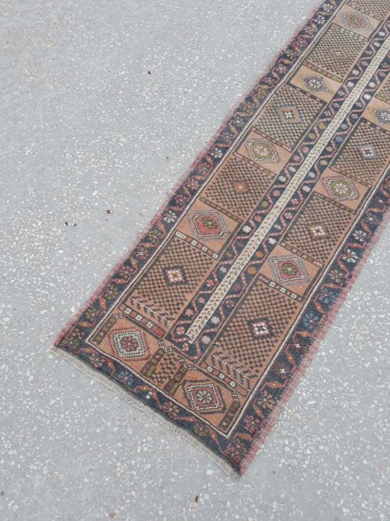 May include: A long, narrow rug with a traditional Persian design. The rug is in shades of brown, pink, and blue, with a central white stripe. The rug has a fringed edge.