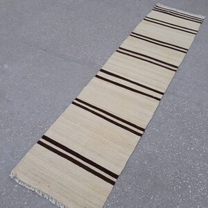 May include: A long, narrow, off-white rug with brown stripes. The rug has a fringe on both ends.