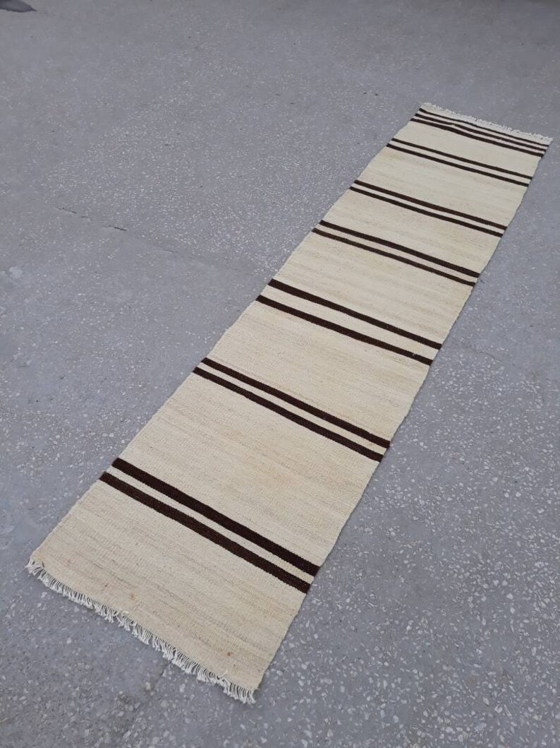 May include: A long, narrow, off-white rug with brown stripes. The rug has a fringe on both ends.