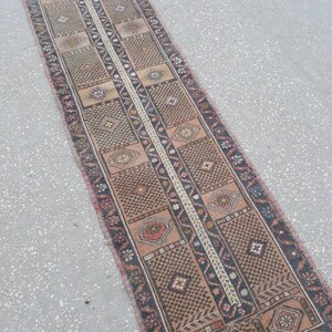 May include: A long, narrow rug with a traditional Persian design. The rug is made of wool and has a brown, beige, and red color scheme. The rug is in good condition and has a vintage look.