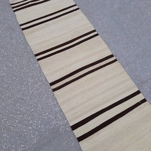 May include: A long, narrow, white rug with brown stripes. The rug is made of a woven material and has a fringed edge.
