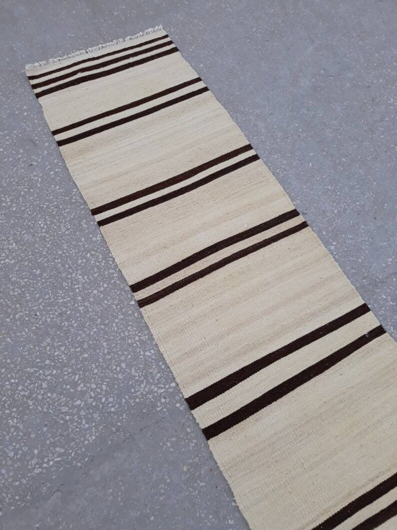 May include: A long, narrow, white rug with brown stripes. The rug is made of a woven material and has a fringed edge.