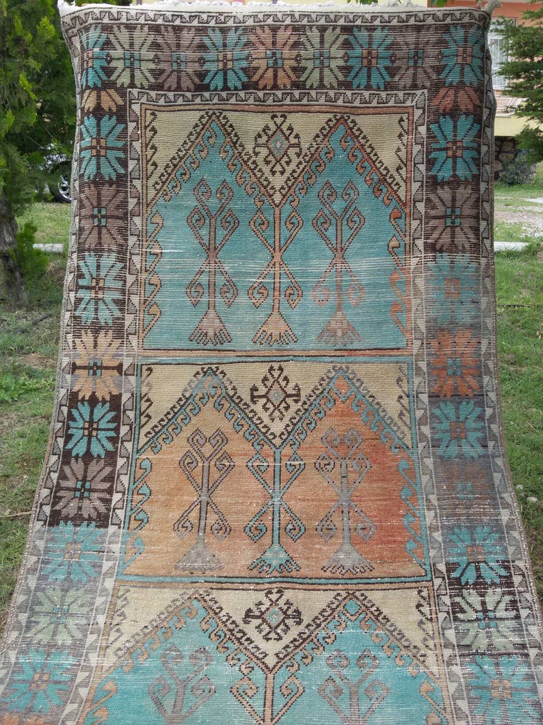 Puede incluir: Una alfombra turca vintage con un patr&oacute;n geom&eacute;trico tradicional en tonos azules, marrones y beige. La alfombra presenta un dise&ntilde;o central de &aacute;rboles estilizados y motivos florales.