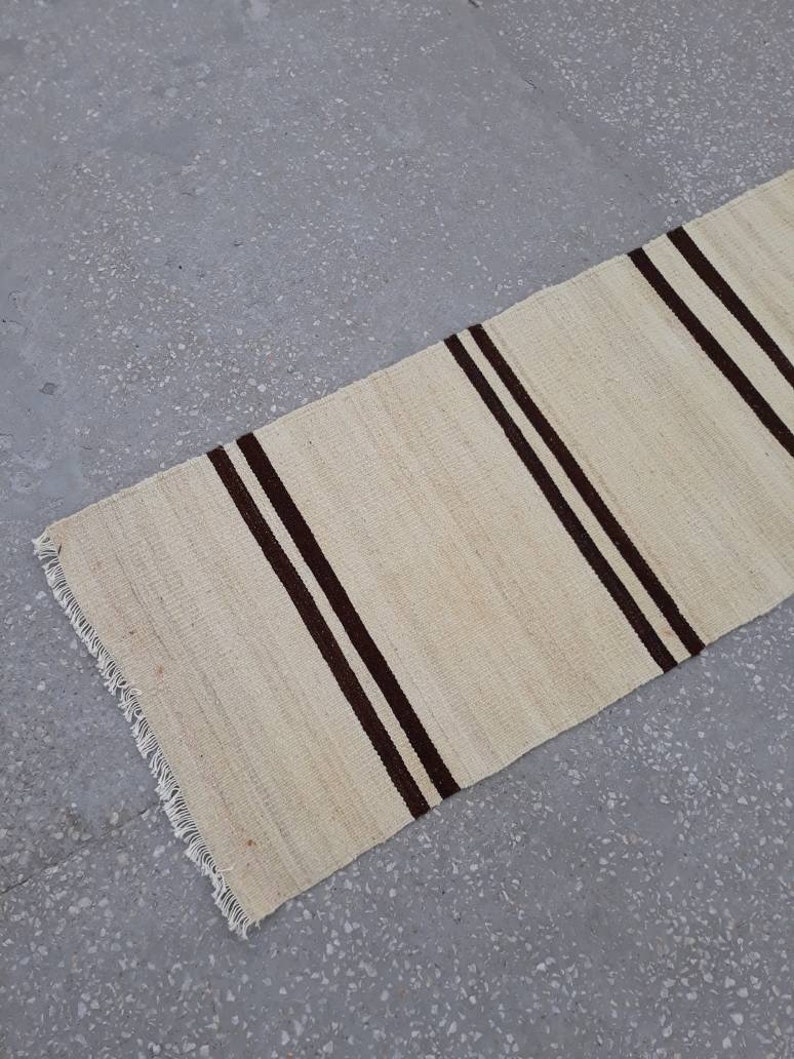 May include: A white and brown striped rug with a fringed edge. The rug is made of natural fibres and has a rustic look.