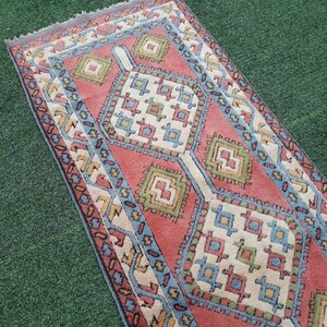 May include: A red, blue, and green patterned rug with a geometric design. The rug has a fringed edge and is laid out on a green surface.