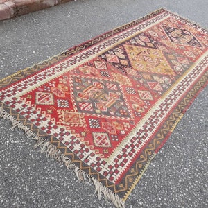 May include: A vintage Turkish rug with a geometric pattern in shades of red, brown, beige, and white. The rug has a fringe border and is woven with intricate details.