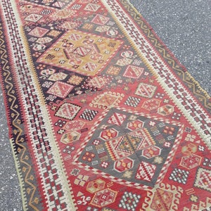 May include: A vintage Turkish rug with a geometric pattern in shades of red, black, white, and gold. The rug has a long, rectangular shape and is woven with intricate details.