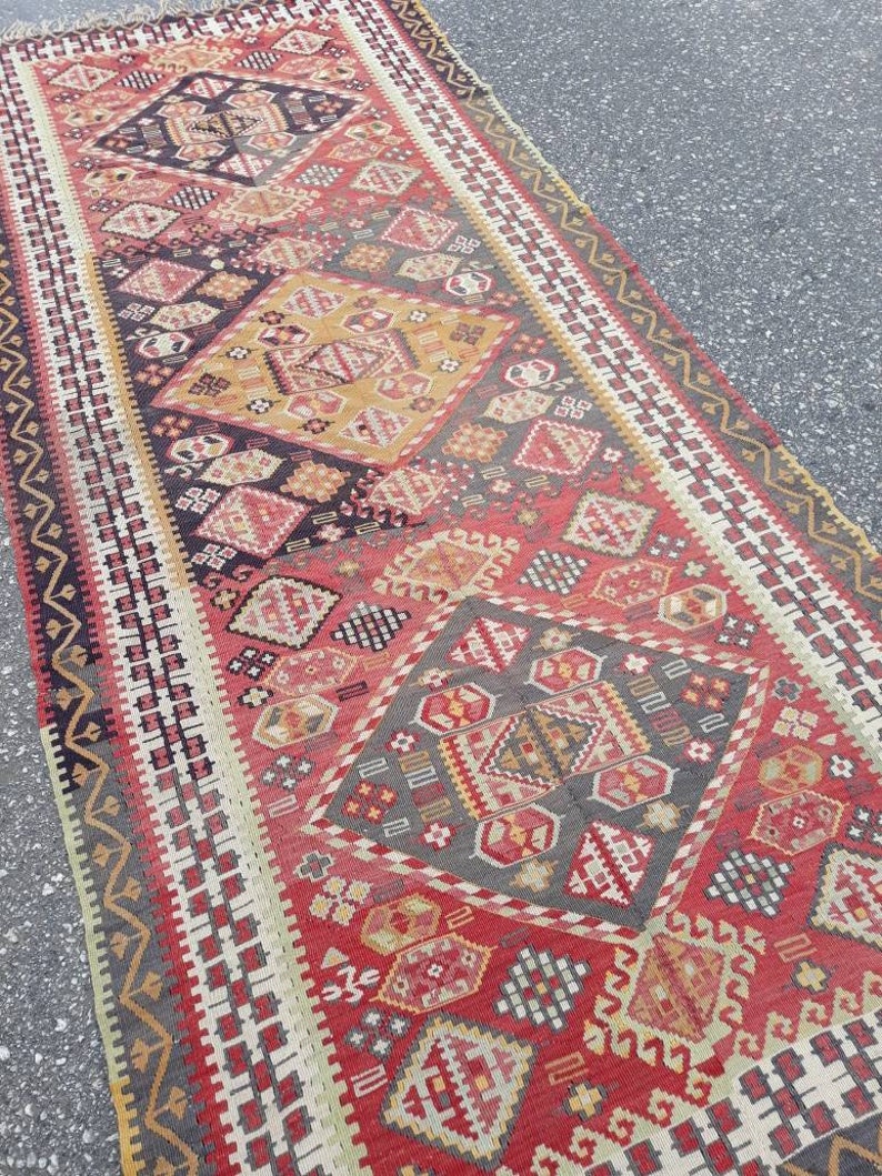 May include: A vintage Turkish rug with a geometric pattern in shades of red, black, white, and gold. The rug has a long, rectangular shape and is woven with intricate details.