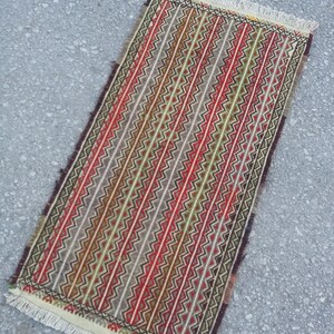 May include: A rectangular rug with a geometric pattern in shades of red, green, brown, and white. The rug has a fringe border.
