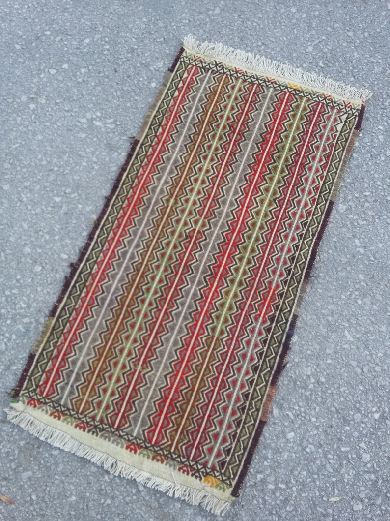 May include: A rectangular rug with a geometric pattern in shades of red, green, brown, and white. The rug has a fringe border.