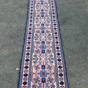 May include: A long, narrow rug with a traditional Persian design. The rug is cream-colored with a pink, blue, and navy blue pattern. The rug has a fringe on both ends.
