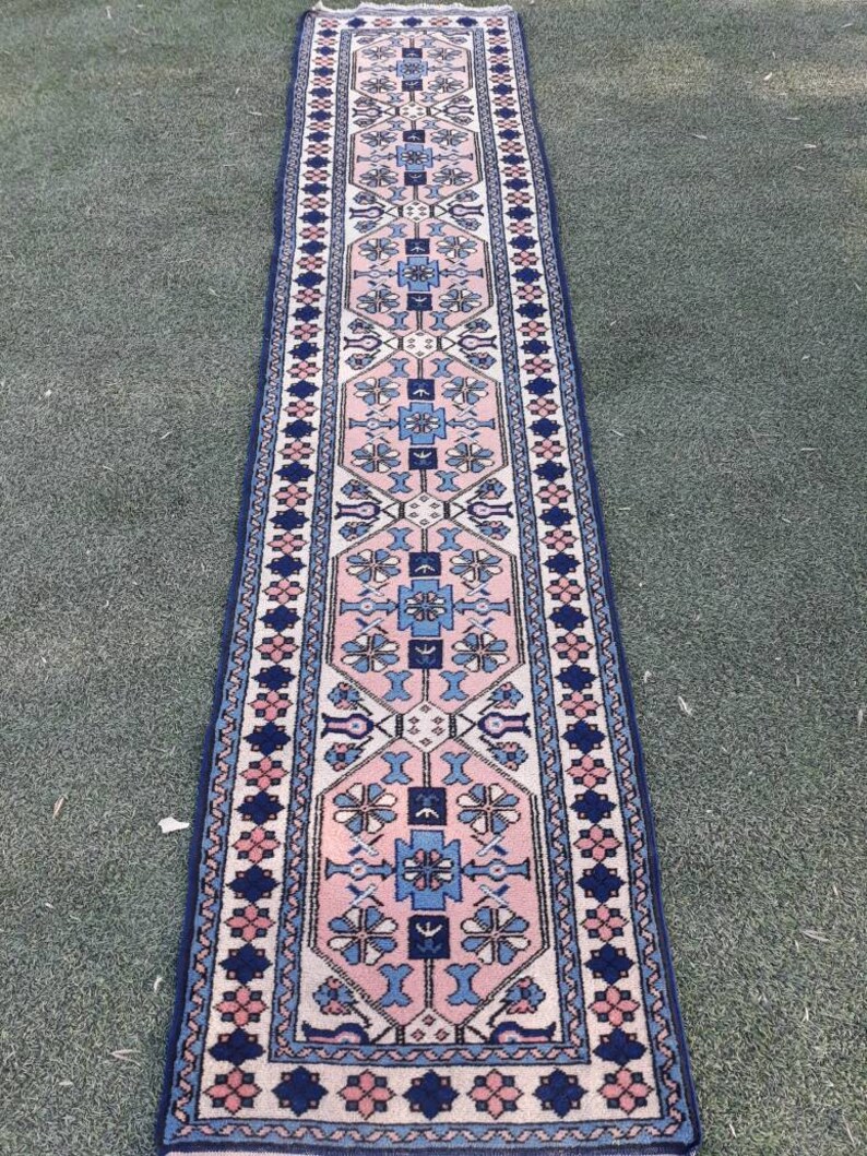 May include: A long, narrow rug with a traditional Persian design. The rug is cream-colored with a pink, blue, and navy blue pattern. The rug has a fringe on both ends.