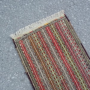 May include: A long, narrow rug with a geometric pattern in shades of red, green, brown, and white. The rug has a fringe on both ends.