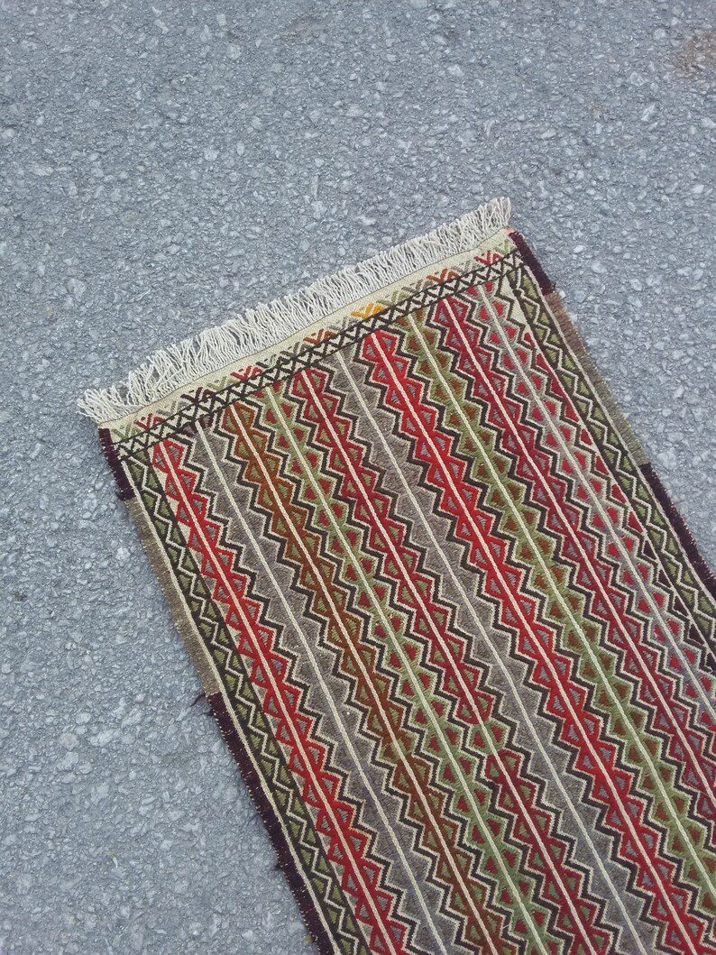 May include: A long, narrow rug with a geometric pattern in shades of red, green, brown, and white. The rug has a fringe on both ends.
