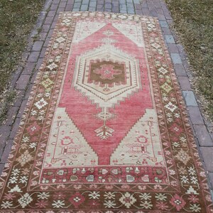 May include: A vintage Turkish runner rug with a pink and cream color scheme. The rug has a geometric pattern with a central medallion and a brown border.
