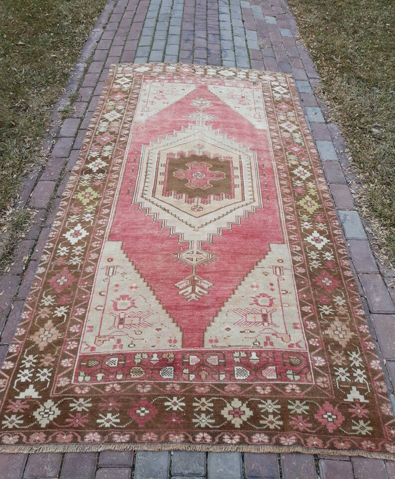 May include: A vintage Turkish runner rug with a pink and cream color scheme. The rug has a geometric pattern with a central medallion and a brown border.