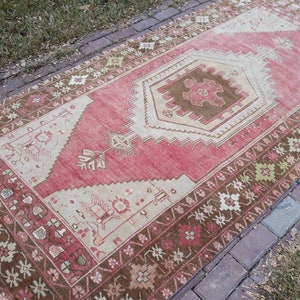 May include: A vintage Turkish runner rug with a pink and beige color scheme. The rug has a geometric pattern with a central medallion and a brown border.