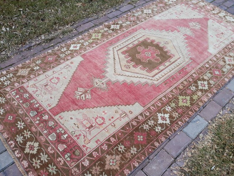 May include: A vintage Turkish runner rug with a pink and beige color scheme. The rug has a geometric pattern with a central medallion and a brown border.