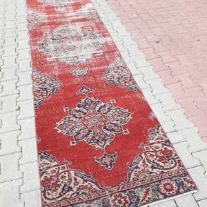 Pode incluir: Um tapete corredor vermelho vintage com um padrão floral desbotado e uma borda azul e branca. O tapete está colocado sobre uma superfície de concreto cinza.