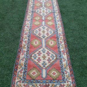 May include: A long, narrow rug with a geometric pattern in shades of pink, blue, green, and white. The rug has a fringe border.