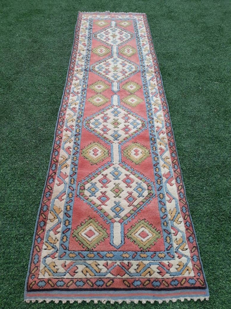 May include: A long, narrow rug with a geometric pattern in shades of pink, blue, green, and white. The rug has a fringe border.