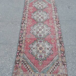 May include: A vintage Turkish runner rug with a pink background and intricate floral patterns in shades of beige, green, and pink. The rug has a fringed edge and is approximately 2 feet wide and 8 feet long.