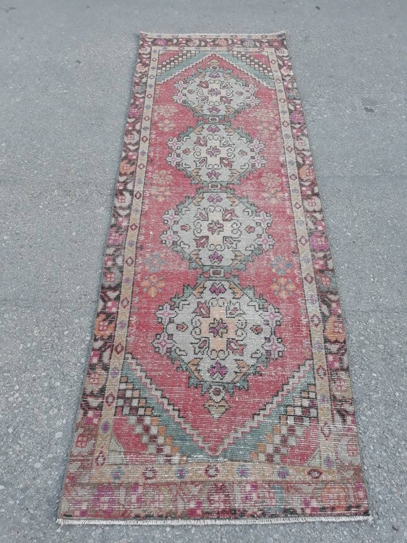 May include: A vintage Turkish runner rug with a pink background and intricate floral patterns in shades of beige, green, and pink. The rug has a fringed edge and is approximately 2 feet wide and 8 feet long.