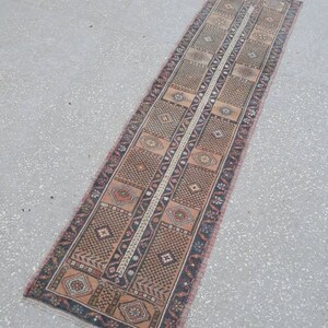 May include: A long, narrow runner rug with a traditional Turkish design. The rug is primarily brown with accents of pink, blue, and white. The rug has a fringe on both ends.