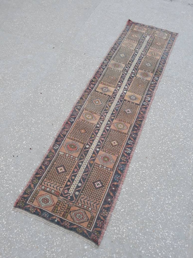 May include: A long, narrow runner rug with a traditional Turkish design. The rug is primarily brown with accents of pink, blue, and white. The rug has a fringe on both ends.