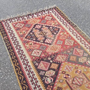 May include: A vintage Turkish rug with a geometric pattern in shades of red, black, white, and gold. The rug features a central diamond shape with intricate details and a border of repeating motifs.