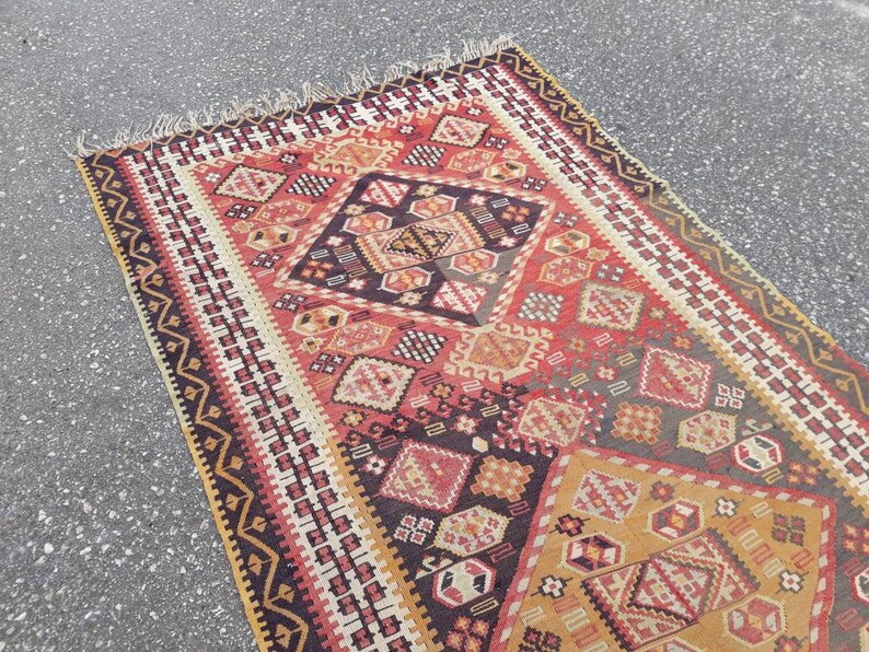 May include: A vintage Turkish rug with a geometric pattern in shades of red, black, white, and gold. The rug features a central diamond shape with intricate details and a border of repeating motifs.