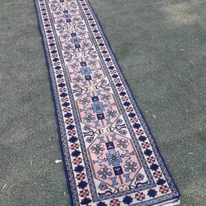 May include: A long, narrow rug with a pink, blue, and white geometric pattern. The rug has a fringe border and is made of wool.
