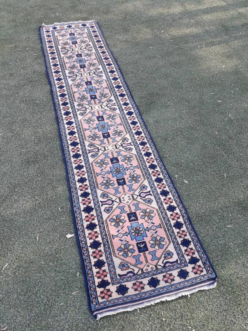 May include: A long, narrow rug with a pink, blue, and white geometric pattern. The rug has a fringe border and is made of wool.