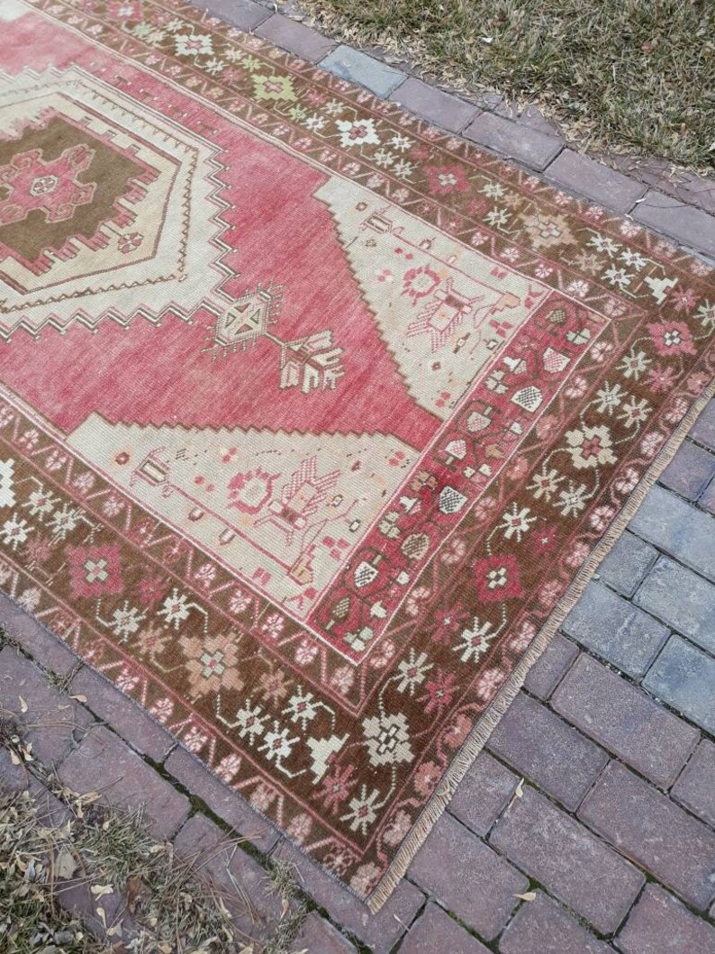 May include: A vintage Turkish runner rug with a red background and a brown and white geometric pattern. The rug has a fringe border and is made of wool.