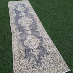 May include: A long, narrow, vintage rug with a faded, cream-colored background and a dark blue, floral pattern. The rug has a fringed edge.