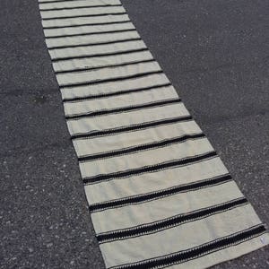 Pu&ograve; includere: Un lungo tappeto da corridoio bianco con strisce nere. Il tappeto &egrave; realizzato in tessuto tessuto e ha un bordo con frange.