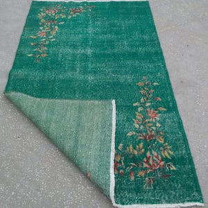 Pode incluir: Um tapete vintage verde com um padr&atilde;o floral em tons de laranja, amarelo e branco. O tapete est&aacute; dobrado ao meio, revelando o verso.