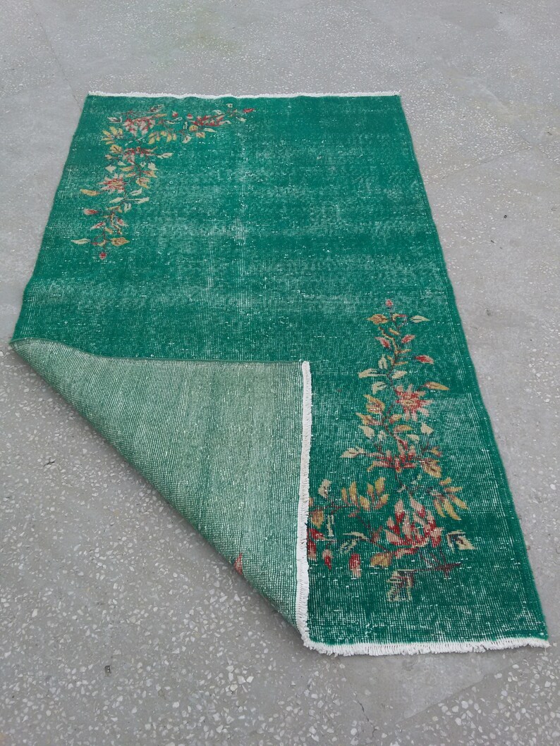 Pode incluir: Um tapete vintage verde com um padr&atilde;o floral em tons de laranja, amarelo e branco. O tapete est&aacute; dobrado ao meio, revelando o verso.