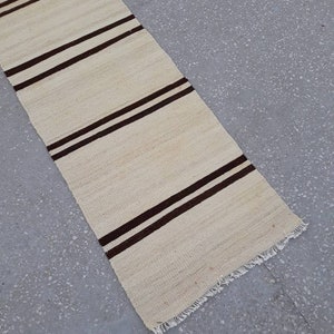 May include: A long, narrow, off-white rug with brown stripes. The rug has a fringed edge.