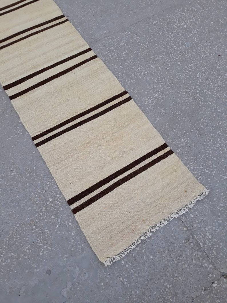 May include: A long, narrow, off-white rug with brown stripes. The rug has a fringed edge.