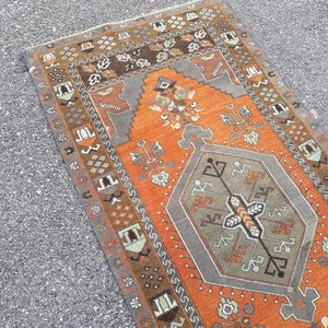 May include: A vintage Turkish runner rug with an orange background and a gray geometric pattern in the center. The rug has a brown, gray, and white border with intricate designs.