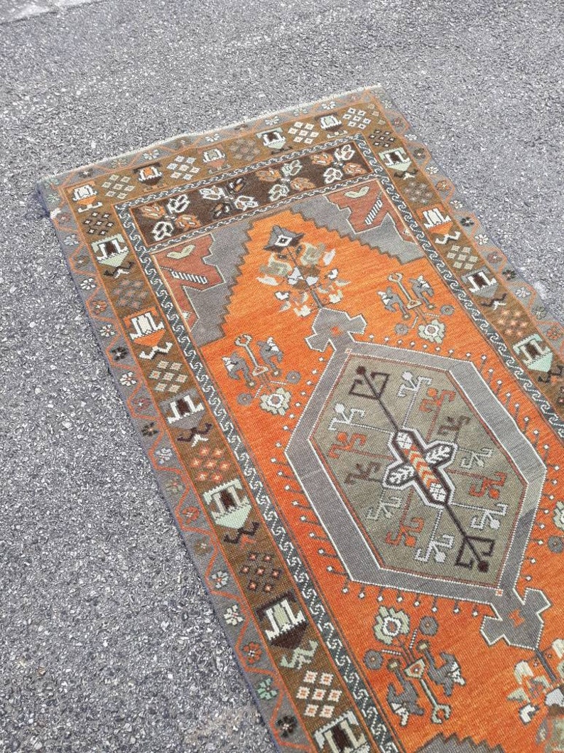 May include: A vintage Turkish runner rug with an orange background and a gray geometric pattern in the center. The rug has a brown, gray, and white border with intricate designs.